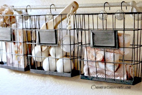 Onemoretimeevents.com. Wire baskets hung on a towel bar using shower curtain hooks.