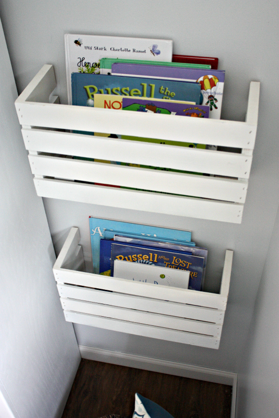 DIY book bins made with a rectangular wooden crate, painted white, cut in half lengthwise and stacked mounted on the wall. The bind hold multiple picture books. 