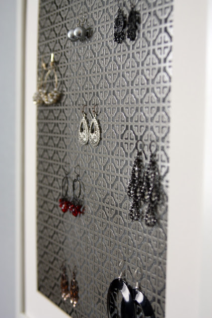 Earring holder made out of a brushed grey metal radiator grate with a geometric pattern and a white frame. Several dangling pairs of earrings hang from grate.