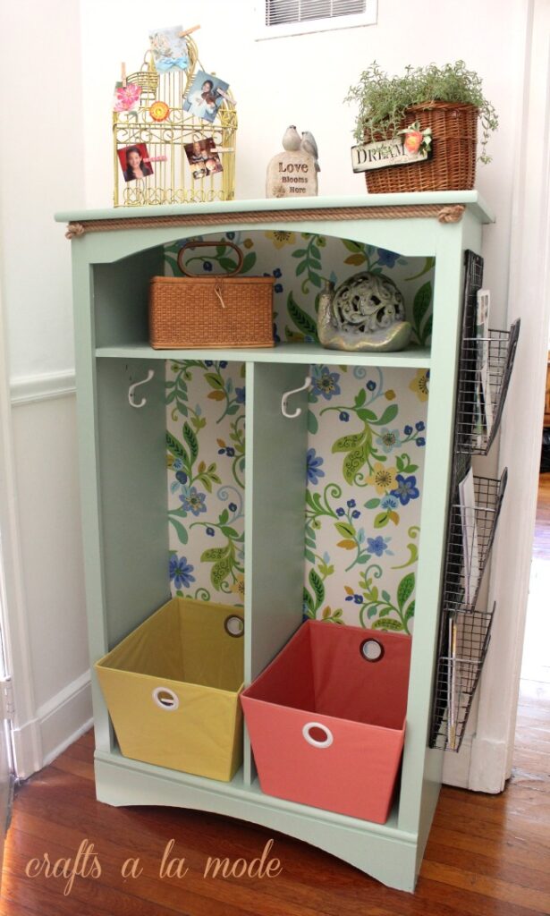 A light green bookshelf reconfigured with top shelf and a vertical slat down the centre with a coat hook installed in each cubby. A large basket, one yellow and one orange are placed at the bottom of each section. A brushed nickel mesh file rack with three sections is attached to side.