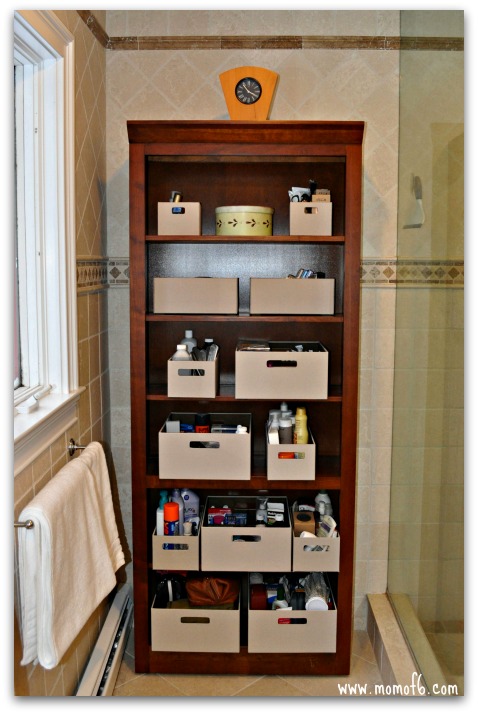 Tall, 6 shelf, dark cherry bookshelf tucked into the corner of a tiled bathroom. Beige fabric baskets are arranged on each shelf filled with toiletries.
