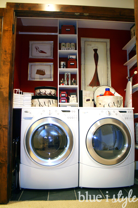 This laundry room maximizes a small space with front-loading machines, vertical shelving, and red accent walls. Stylish bins and artwork add elegance, while labeled storage and baskets provide function. A balance of utility and flair, the space is cleverly organized to make laundry efficient within a closet. blueistyle